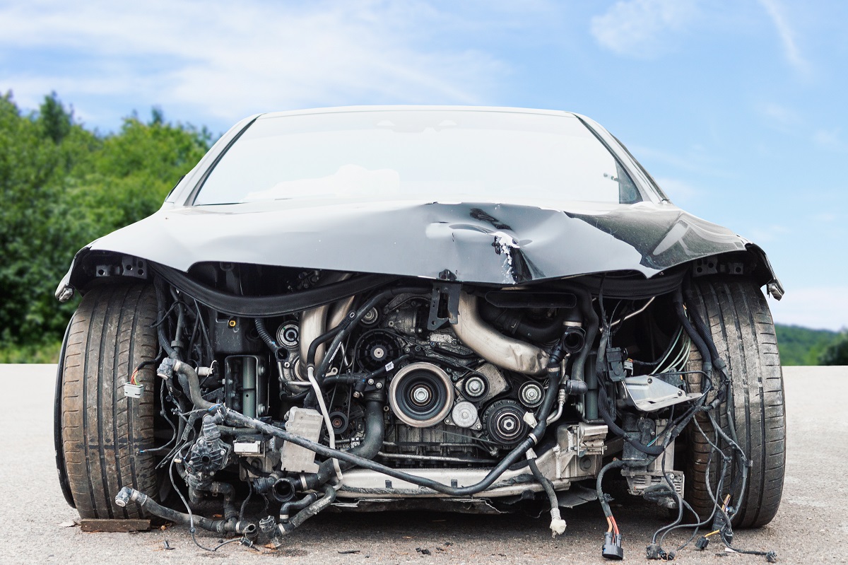 A gray car has a crumpled hood and an exposed engine. The grill and front bumper are missing.