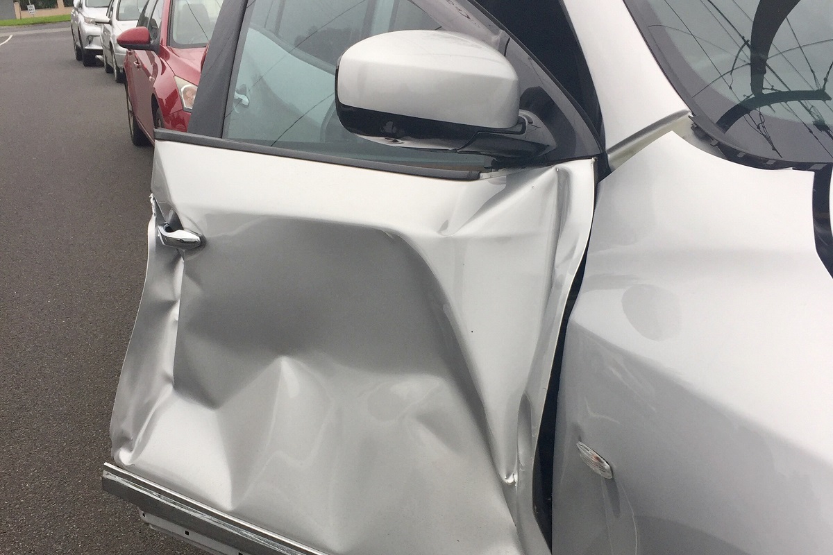 A silver car parked on the side of the road with a dented door that is slightly open.